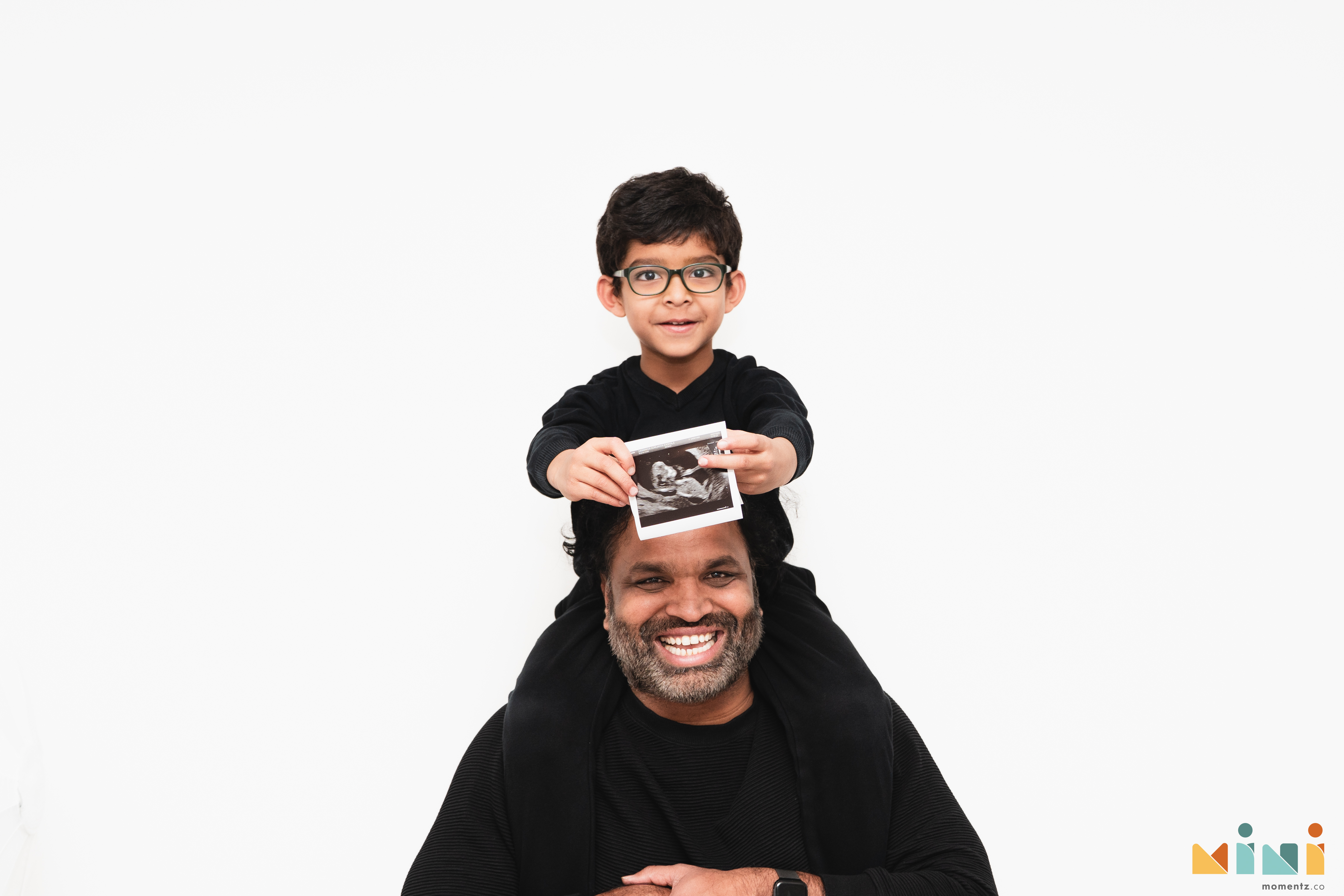 Kiwi sitting on his father Chaithanya's shoulders, proudly holding up his sibling's ultrasound scan photo during the MiniMomentz maternity session in Scotland